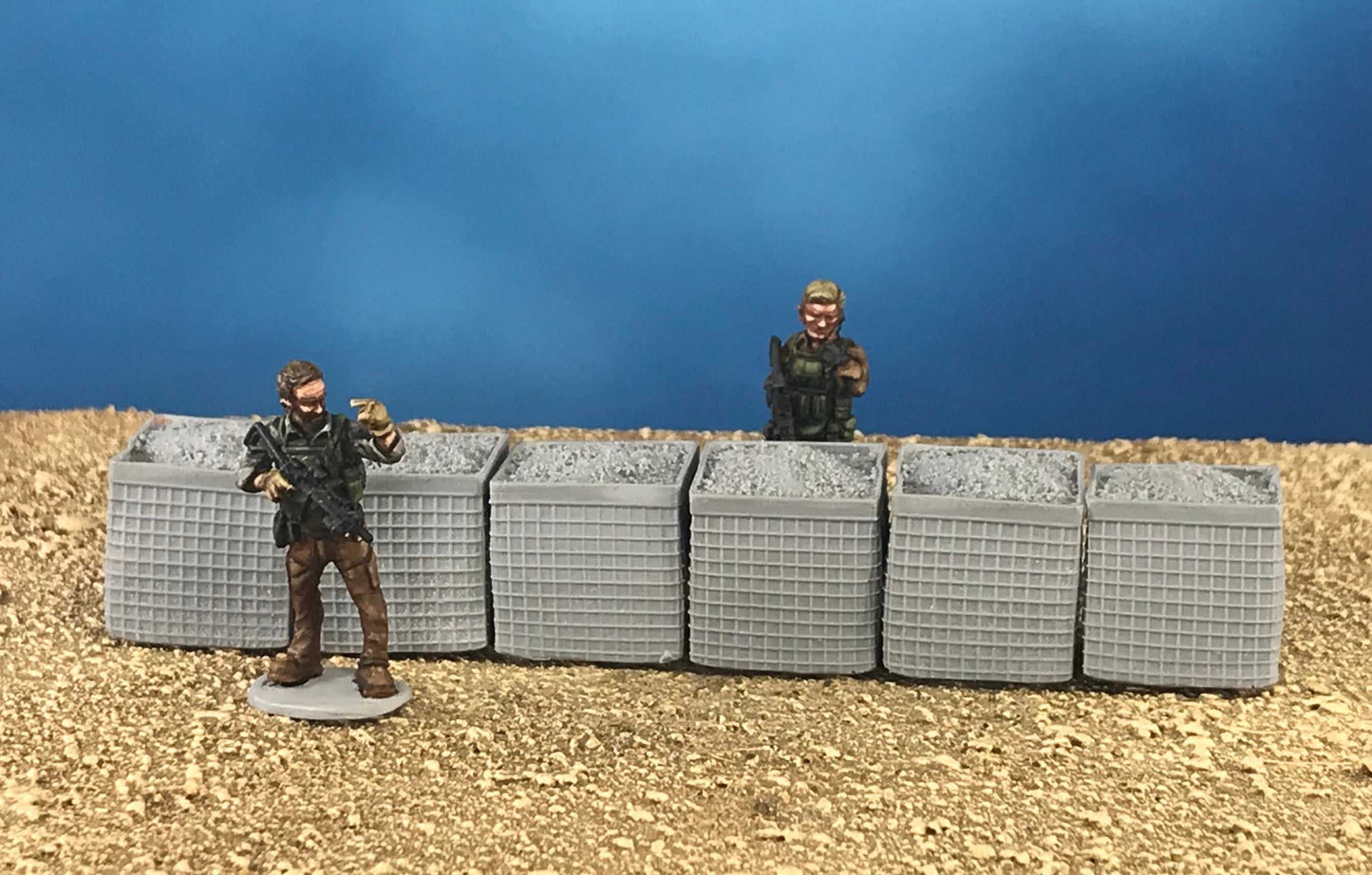 Set of six 28mm modern hesco barriers on sandy terrain with two miniature figures behind and beside them